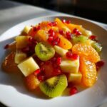 A vibrant serving of winter fruit salad featuring bright orange segments, kiwi slices, apple cubes, and ruby red pomegranate seeds.