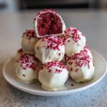 A stack of white chocolate covered red velvet cake pops topped with pink and red sprinkles, one is cut open.