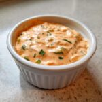 Close-up of creamy, orange-hued remoulade sauce speckled with green herbs in a white dipping bowl.