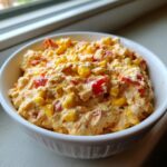 Close-up of a creamy, orange-tinted boat dip filled with visible kernels of yellow corn and diced red peppers in a white bowl.