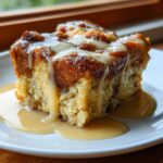A moist, square slice of bread pudding topped with a thick vanilla glaze and sitting in a pool of caramel sauce.