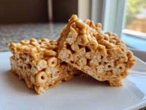 Two squares of homemade cheerio bars, rich with peanut butter coating, resting on a white plate.