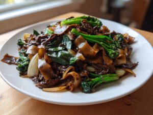 A close-up of a generous serving of amazing thai drunken noodles featuring wide rice noodles, dark sauce, beef, and bright green broccoli.