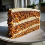 A tall, three-layer slice of carrot cake cheesecake topped with thick cream cheese frosting.