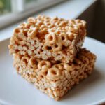 Close-up of two stacked, gooey cheerio bars made with cereal rings and a sweet binder, resting on a white plate.