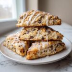 A stack of four triangular chocolate chip scones drizzled with white vanilla glaze, sitting on a white plate.