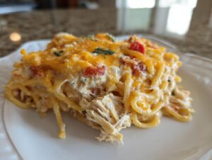 A serving of creamy chicken spaghetti topped with melted cheddar cheese and red peppers on a white plate.