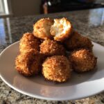 A pile of golden brown, crispy fried goat cheese balls on a white plate, one is broken open showing the soft interior.