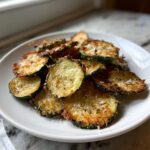 A pile of golden, crispy zucchini chips topped with Parmesan cheese and pepper served on a white plate.