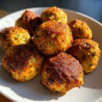 A close-up of several golden brown, crispy stuffing balls piled on a white plate, beautifully lit by sunlight.