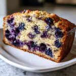 A close-up view of a thick, moist slice of blueberry banana bread on a white plate.