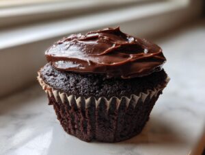 A single, moist chocolate cupcake topped with thick, glossy chocolate frosting, sitting on a light marble surface.