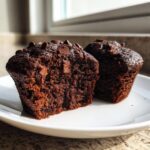 Close-up of two rich, dark chocolate muffins, one cut open, topped with chocolate chips on a white plate.
