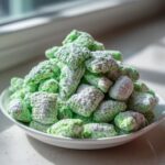 A close-up of bright green pistachio puppy chow heavily dusted with powdered sugar, piled high in a white bowl.