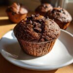 Close-up of a freshly baked, dark chocolate chip muffin topped with melted chips on a white plate.