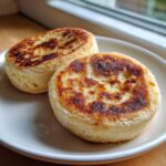 Two golden-brown sourdough english muffins with characteristic nooks and crannies, resting on a white plate.