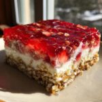 A close-up, sunlit slice of strawberry pretzel salad showing three distinct layers: pretzel crust, cream cheese filling, and bright red strawberry gelatin topping.