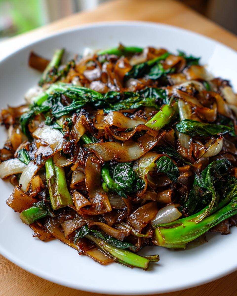 Close-up of glossy, dark brown thai drunken noodles mixed with vibrant green Chinese broccoli and onions on a white plate.