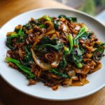 A close-up of a plate piled high with savory thai drunken noodles, dark sauce, onions, and vibrant green Chinese broccoli.