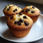 Close-up of three freshly baked mini chocolate chip muffins with dark chocolate chips on top, resting on a white plate.