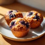 Three golden-brown blueberry cottage cheese muffins resting on a light-colored plate near a window.