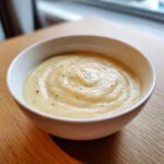 A close-up of a white bowl filled with smooth, creamy garlic soup, lightly seasoned with black pepper.