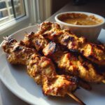 Close-up of grilled chicken satay skewers on a white plate next to a bowl of peanut sauce.