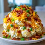 A mound of creamy loaded baked potato salad topped generously with shredded cheddar, crispy bacon bits, and green onions.