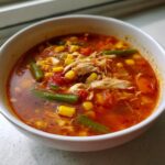 Close-up of a bowl of rich, tomato-based maryland crab soup filled with shredded chicken, corn, and green beans.