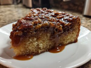 A close-up of a moist slice of pecan upside down cake dripping with rich caramel sauce on a white plate.
