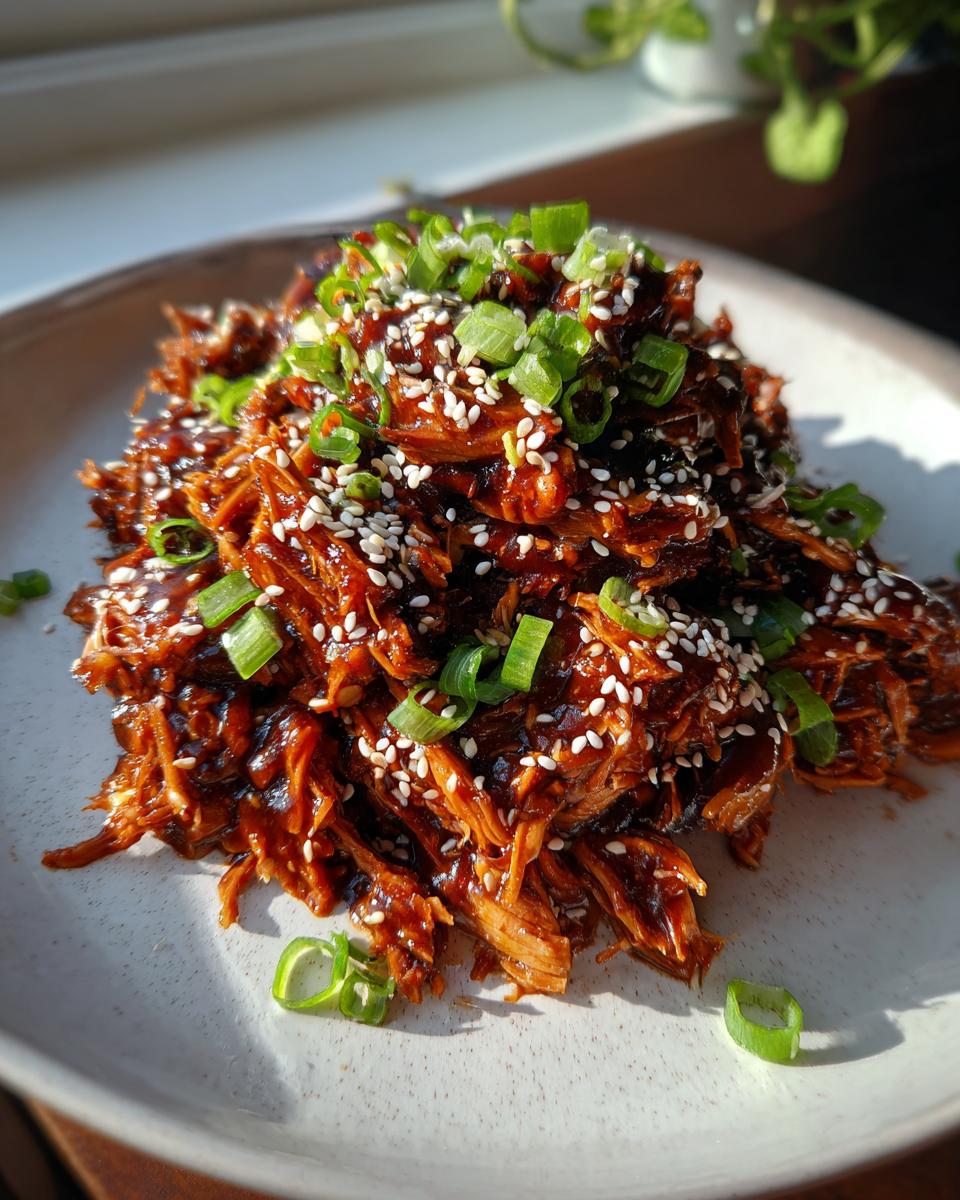 A mound of saucy, shredded crockpot teriyaki chicken topped with white sesame seeds and sliced green onions.