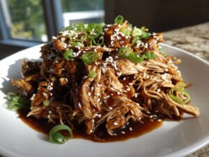 Close-up of shredded crockpot teriyaki chicken drizzled with sauce, topped with sesame seeds and green onions.