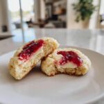Two halves of a soft strawberry cheesecake cookie showing the creamy filling and bright red strawberry jam topping.