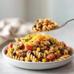 A fork lifting a bite of creamy taco pasta salad mixed with ground beef, corn, and black beans from a white bowl.
